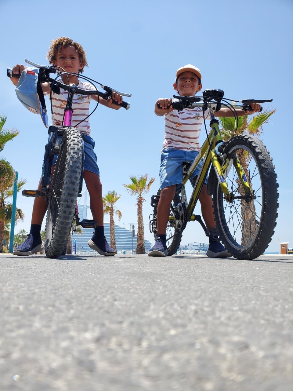 Two young riders on bikes on a waterfront path, with Dubai's skyline behind them